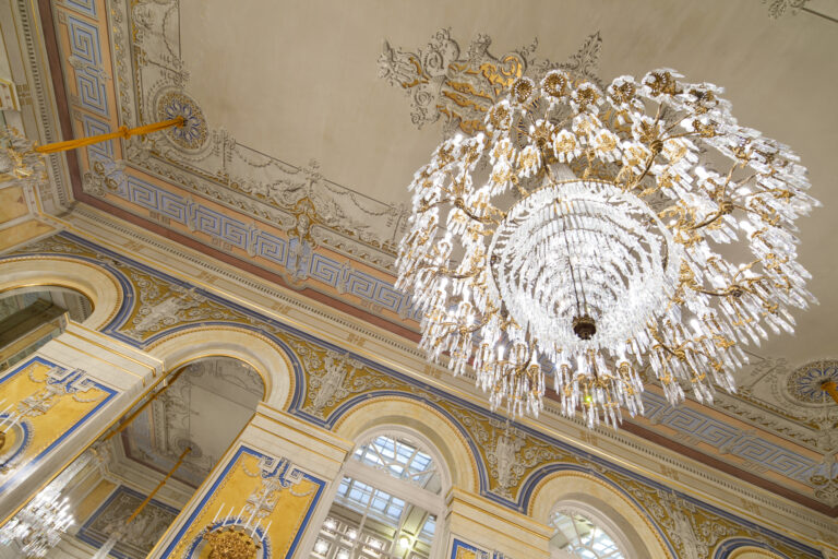 Stor kristallkrona hänger från ett utsmyckat tak.