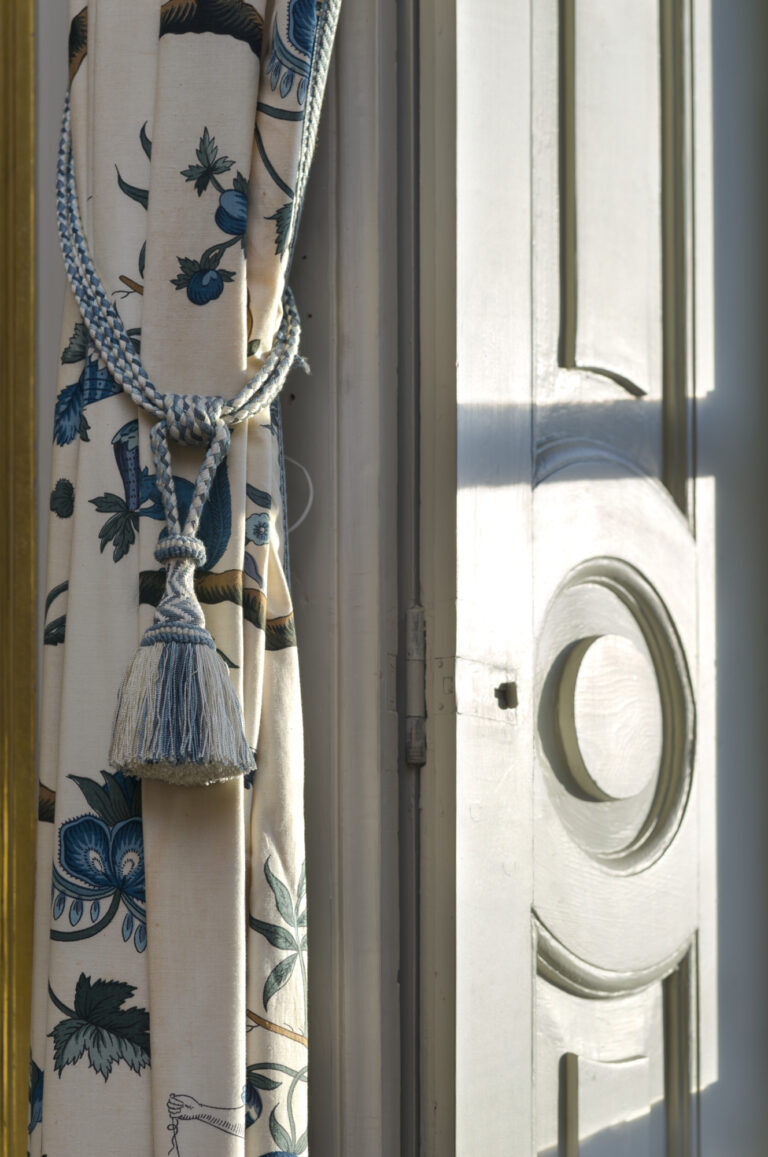 Inzoomad bild på en fönsterlucka i trä och en blåblommig gardin.