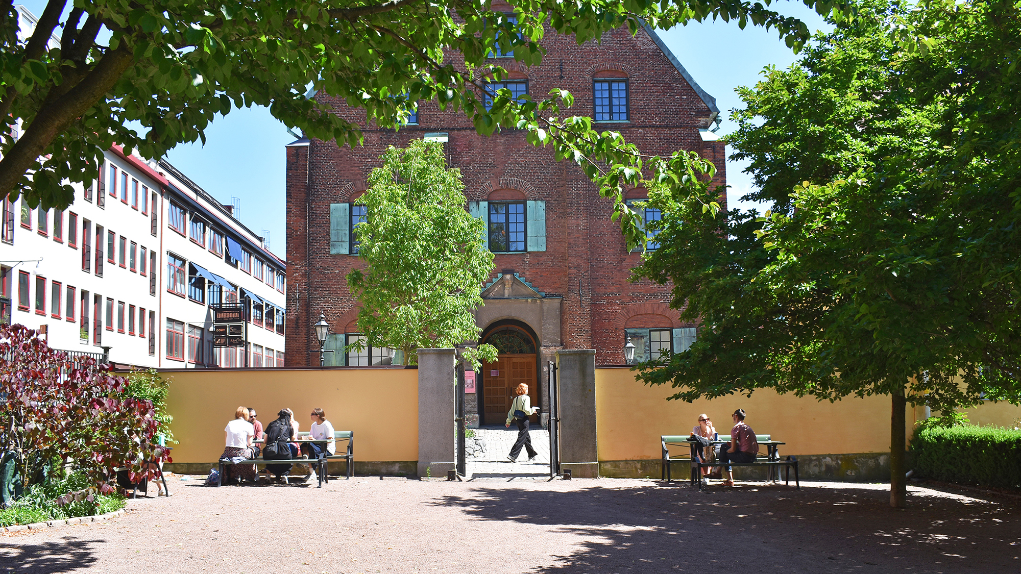 Gröna träd ramar in en rött tegelhus och en park där människor sitter vid bänkar.